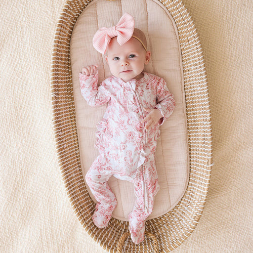 Baby in a floral onesie with a pink bow in a woven basket on a beige background
