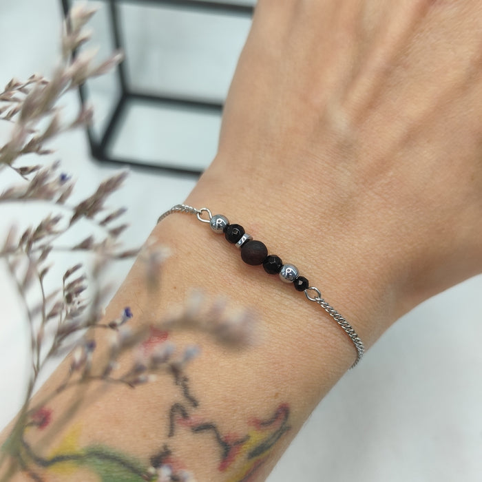 Armband aus Räuchereiche mit Holzperlen und silberfarbener Kette am Handgelenk getragen, im Hintergrund eine Ecke Glaskasten und Strauch im Vordergrund