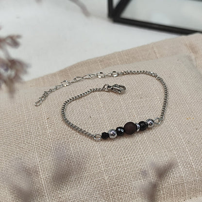 Armband aus Räuchereiche mit Holzperlen und silberfarbener Kette auf einem braunen Webare-Kissen, im Hintergrund eine Ecke Glaskasten und Strauch im Vordergrund