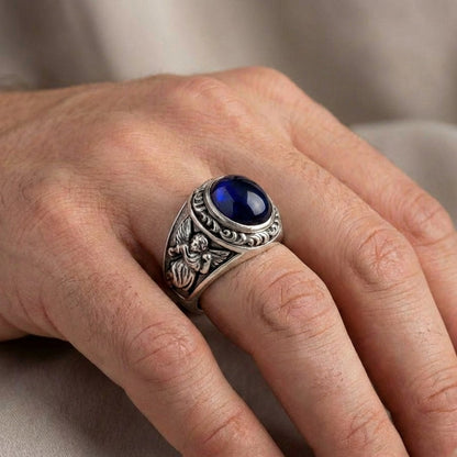 Silver men's ring with oval blue cabochon stone and engraved cupid angel design worn on hand, featuring ornate baroque scrollwork detailing on the band