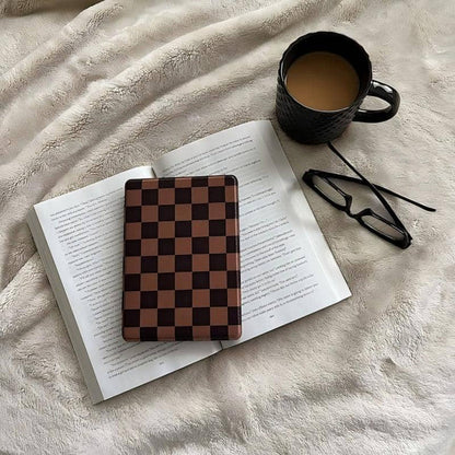 Open book with a checkered kindle case object on top, next to a black mug and glasses on a soft surface.