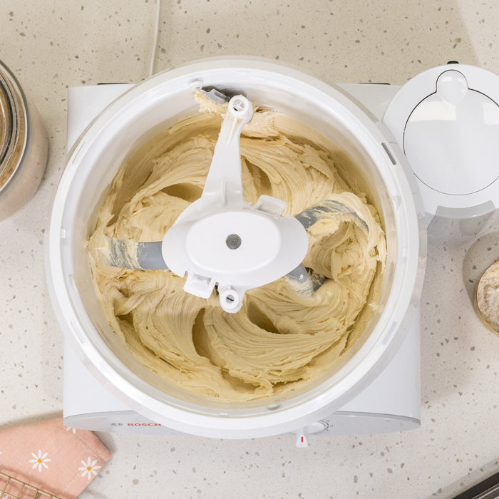 Bosch white universal plus mixer with NutriMill bowl scraper and pro line cookie paddles with metal whip drive on white speckled counter