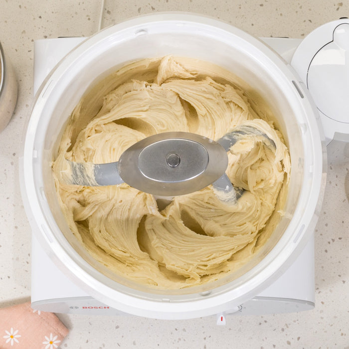Bosch white universal plus mixer mixing white dough with pro line cookie paddles and metal whip drive on white speckled counter