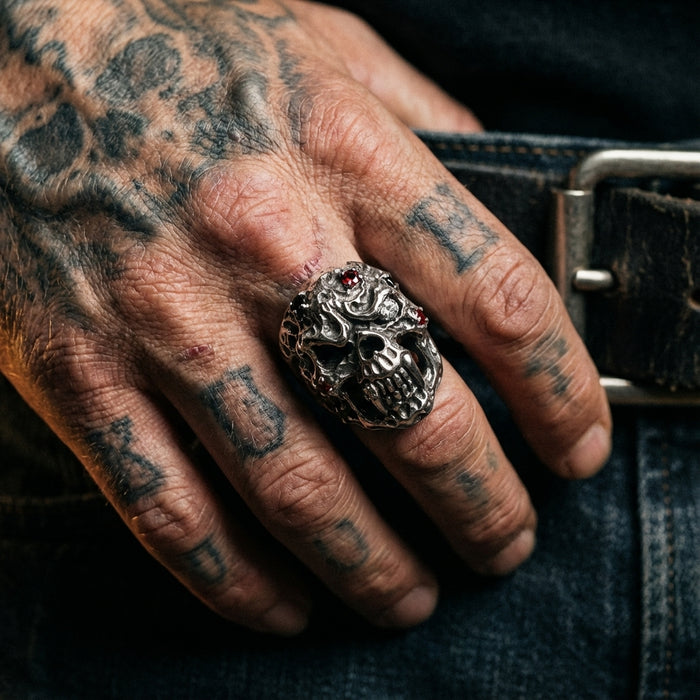 Close-up of a heavily tattooed hand wearing a large sterling silver vampire skull ring adorned with diamonds and red gemstones, resting against blue denim jeans and a leather belt.