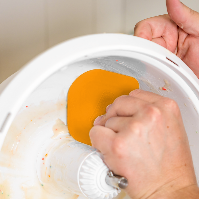 Hand using a NutriMill orange easy scrape bowl scraper to scrape funfetti batter from inside a mixer bowl