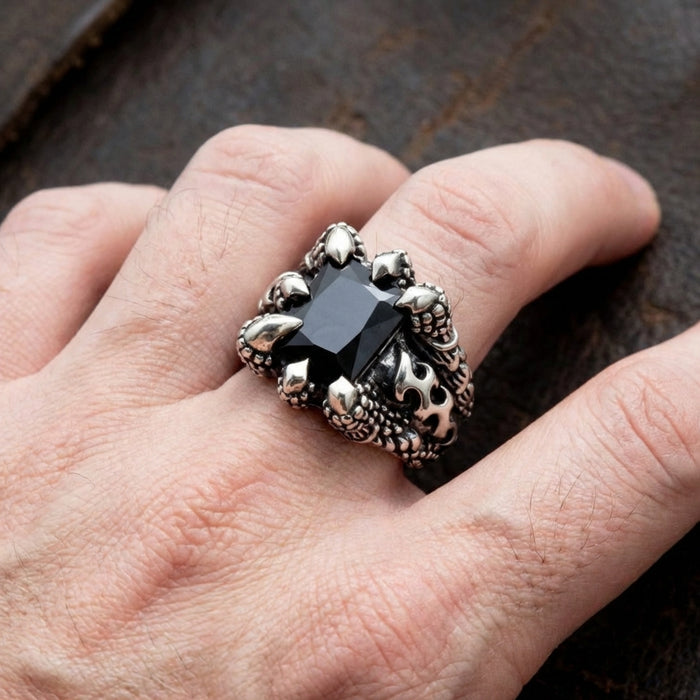 Silver gothic claw ring featuring a large rectangular black onyx gemstone held by dragon claw prongs with fleur-de-lis detailing, worn on a man's finger against a dark rustic background