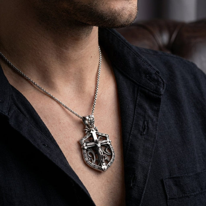 Man wearing a sterling silver Gothic crucifix shield pendant necklace with ornate filigree detailing and stone accents on a box chain, styled with an open dark navy shirt