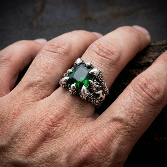 Gothic silver ring with large emerald green gemstone held by claw prongs and dragon scale detailing worn on a man's finger against dark background