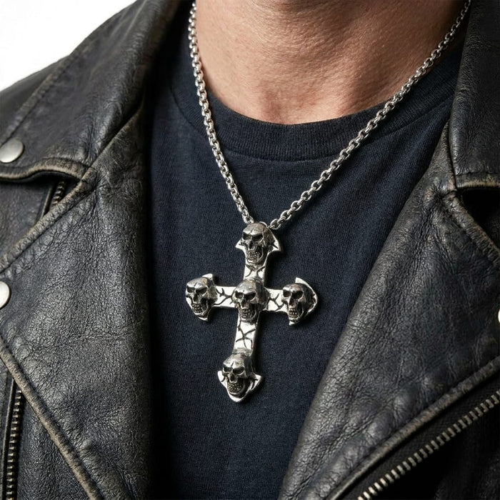 Close-up of a man wearing a distressed black leather jacket and black t-shirt, modeling a heavy silver cross pendant necklace featuring multiple detailed skulls in a gothic biker style.