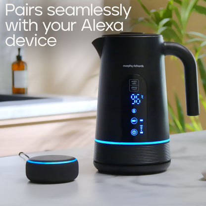 A black Morphy Richards electric kettle with a digital display and blue LED lighting, shown alongside a matching smart control dock on a kitchen counter. The image includes a text overlay that reads 'Pairs seamlessly with your Alexa device'.