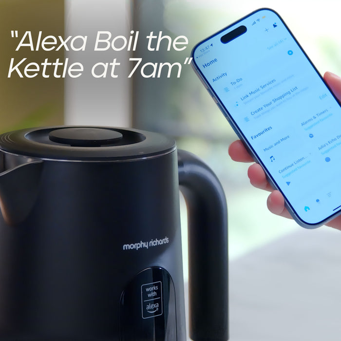 A black Morphy Richards electric kettle with Alexa enabled, shown next to a hand holding a smartphone displaying the Alexa app with a scheduled wake-up reminder text overlay.