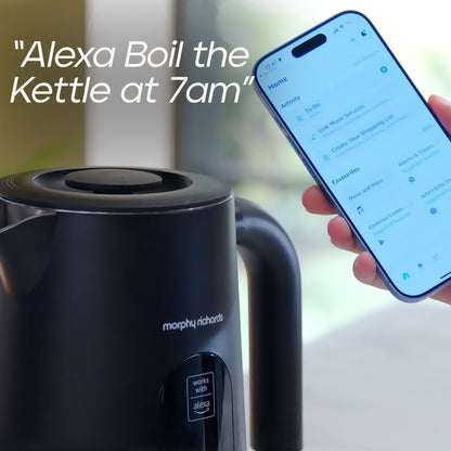 A black Morphy Richards electric kettle with Alexa enabled, shown next to a hand holding a smartphone displaying the Alexa app with a scheduled wake-up reminder text overlay.