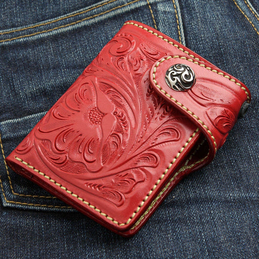 A hand-carved red leather biker wallet with a floral pattern and decorative silver snap, resting on blue jeans.