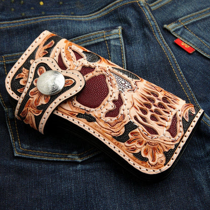 A classic long biker wallet with a finely hand-carved leather skull, shown resting on a dark denim background.