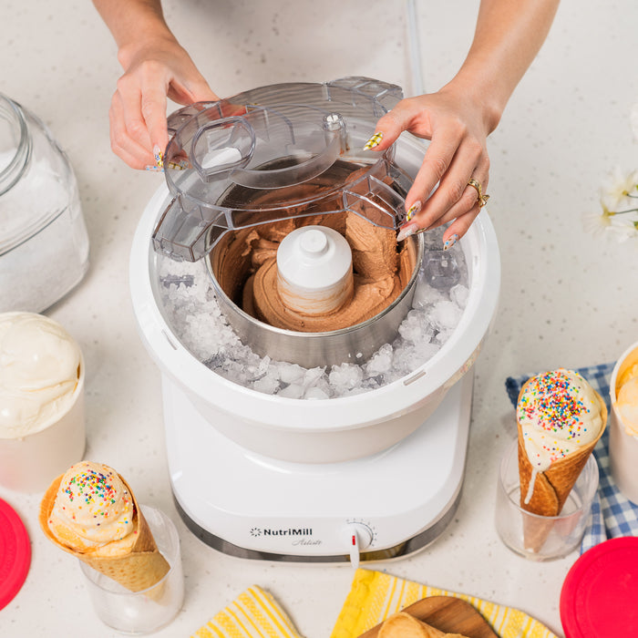 Lifestyle photo of hands using a white ice cream maker with ice cream cones and dishes nearby