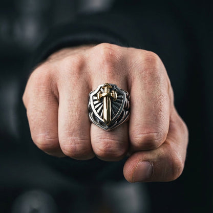 Close-up of a hand wearing a two-tone silver and gold knight shield ring featuring a raised gold sword emblem, photographed in moody lighting with a blurred dark background.