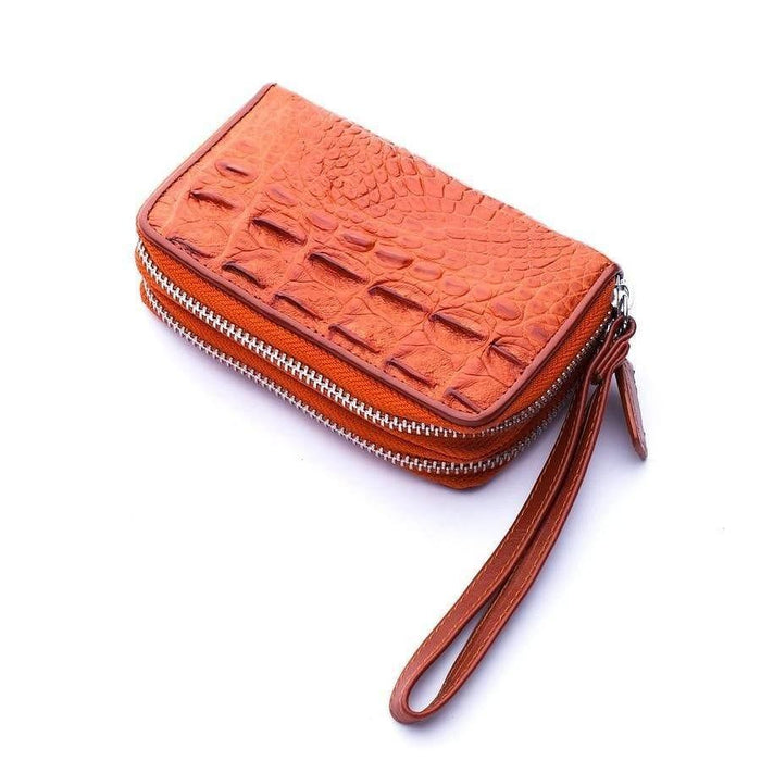 Three-quarter overhead shot of orange-toned crocodile leather wristlet wallet with silver zipper and leather strap on white surface