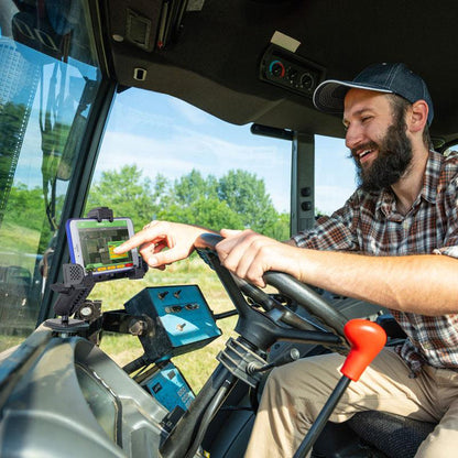 A bearded man in a plaid shirt and cap smiles while using a touchscreen device on the LockVise™ Locking Phone Mount with 2.75 Arm and Metal AMPS Drill Base, installed near the tractors steering wheel. Greenery and blue sky are visible outside.