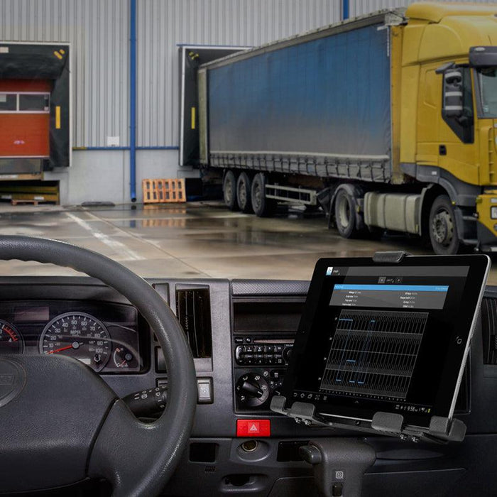 View from inside a truck cab showing the LockVise™ Metal Locking Tablet Mount with Drill Base and Magnetic Lightning Charge Cable displaying a graph, with parked trucks and a loading dock visible through the windshield.
