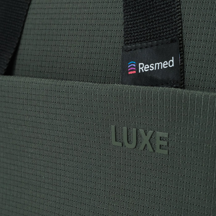 Luxe Tote bag, Close-up of a dark olive green textured fabric bag with a black webbing strap and a black Resmed brand label sewn onto it. The word 'LUXE' is debossed on the front.