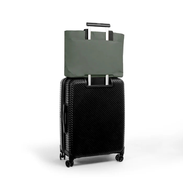 An olive green tote bag with black handles is shown hanging from the telescoping handle of a black hard-shell spinner suitcase on a clean white background.