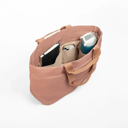 Luxe tote bag A dusty rose canvas tote bag shown open from an overhead angle, filled with a black notebook, a beige phone case, and a teal insulated tumbler, on a clean white background.