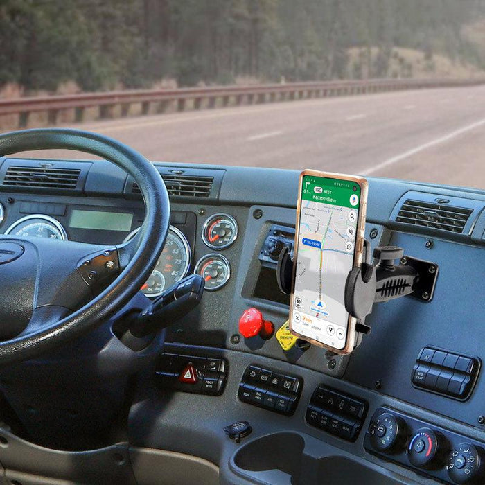 A Drill-Base Mega Grip™ Phone Mount secures a smartphone with navigation on a truck dashboard; the adjustable arm holds the phone near the steering wheel and control panel, with a highway scene visible through the windshield.