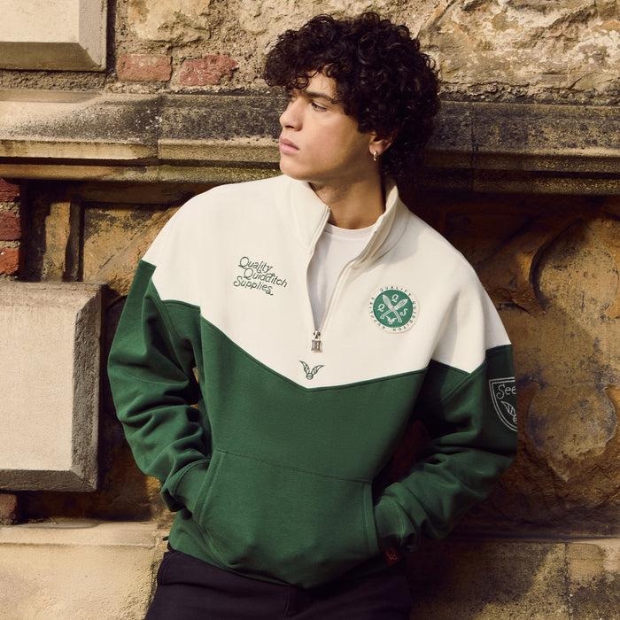 Person wearing a green and white Quidditch sweatshirt with logos, leaning against a stone wall.