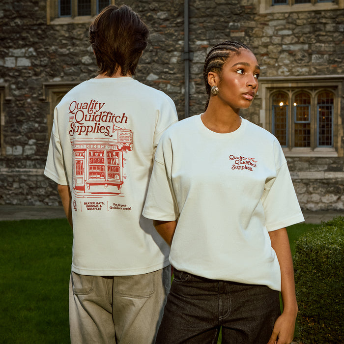 Two people wearing 'Quality Quidditch Supplies' t-shirts in front of a stone building.