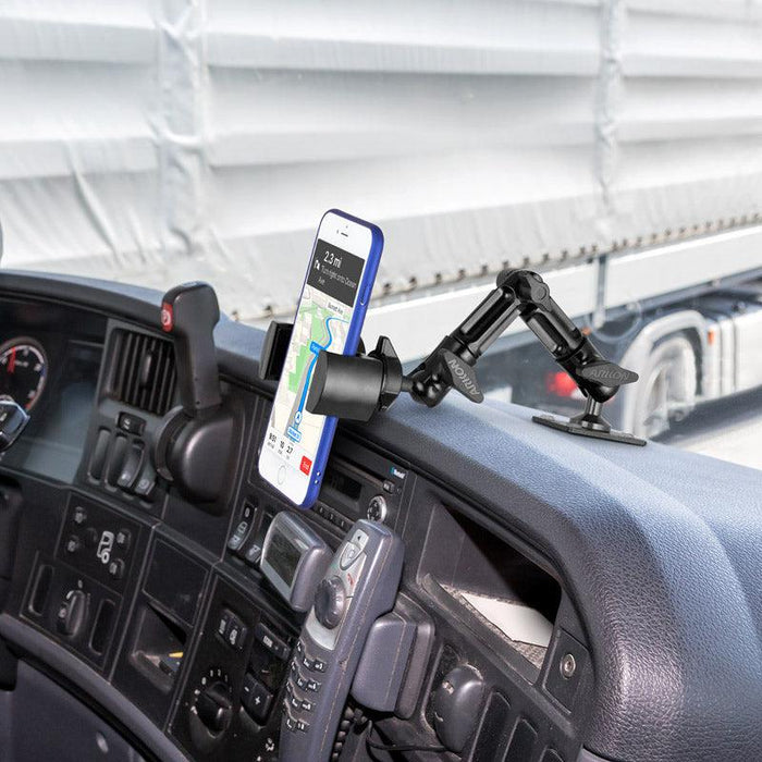 A smartphone on a truck dashboard displays a navigation map, secured by the RoadVise Heavy-Duty Multi-Angle Phone Mount (4-Hole AMPS Compatible), with the steering wheel and window showing a large truck parked outside.