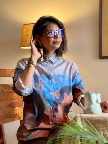 Woman wearing the Shakuntala shirt, seated by a lamp with a mug, hand at her hair