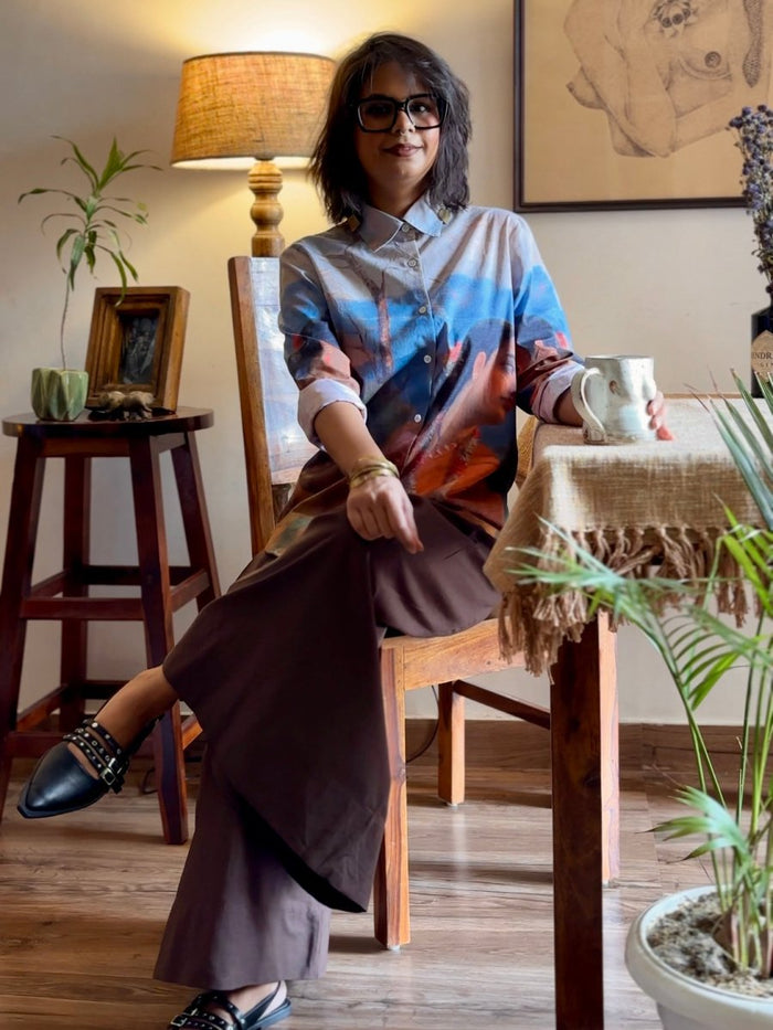 Woman wearing the Shakuntala shirt with brown trousers, seated on a wooden chair, full length view