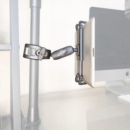 A Slim-Grip® Ultra Phone or Midsize Tablet Clamp Mount with Security Knob holds a tablet on a vertical metal pole beside a desk with a computer monitor in the background.