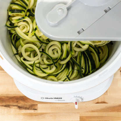 A NutriMill artiste mixer using a NutriMill sprializer attachment to cut zucchini on a wooden counter