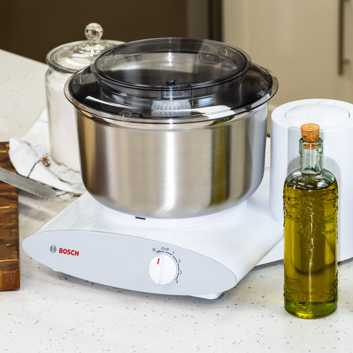 Bosch White Universal Plus Mixer with stainless steel bowl on white counter next to oil.