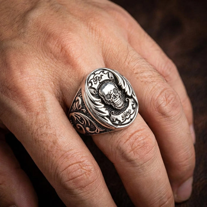 Close-up of a man's hand wearing a chunky silver Por Vida skull biker ring on his middle finger, featuring a central skull design, engraved banners, and ornate gothic filigree detailing on the band.