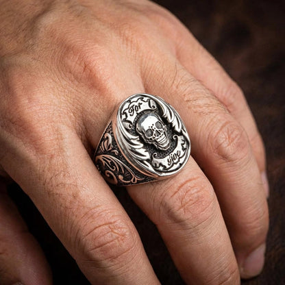 Close-up of a man's hand wearing a chunky silver Por Vida skull biker ring on his middle finger, featuring a central skull design, engraved banners, and ornate gothic filigree detailing on the band.