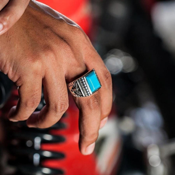 A man wearing a large sterling silver Turquoise eagle ring on his finger, with a blurred red and black background.