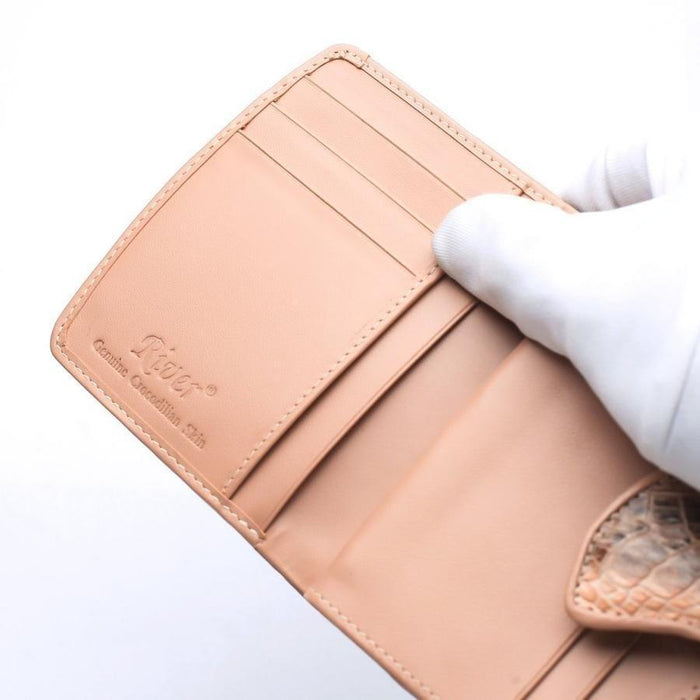 Gloved hand holding open wallet showing River Genuine Crocodilian Skin embossed logo, card slots, and smooth peach leather lining