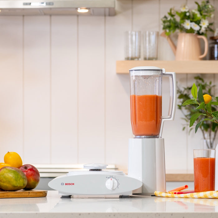 Blender with a red drink inside on a white kitchen counter next to fruit with a kitchen in background