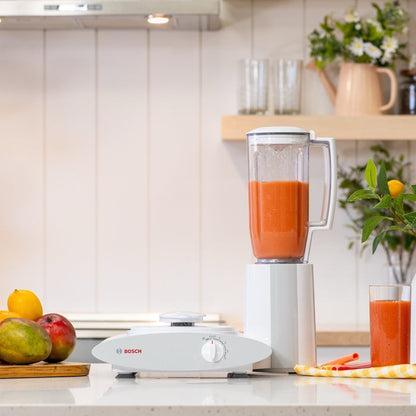 Blender with a red drink inside on a white kitchen counter next to fruit with a kitchen in background