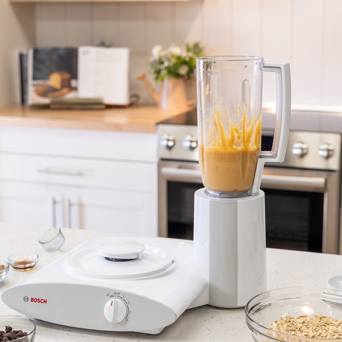 Bosch blender attachment with yellow-orange drink inside, sitting on white counter with kitchen in background