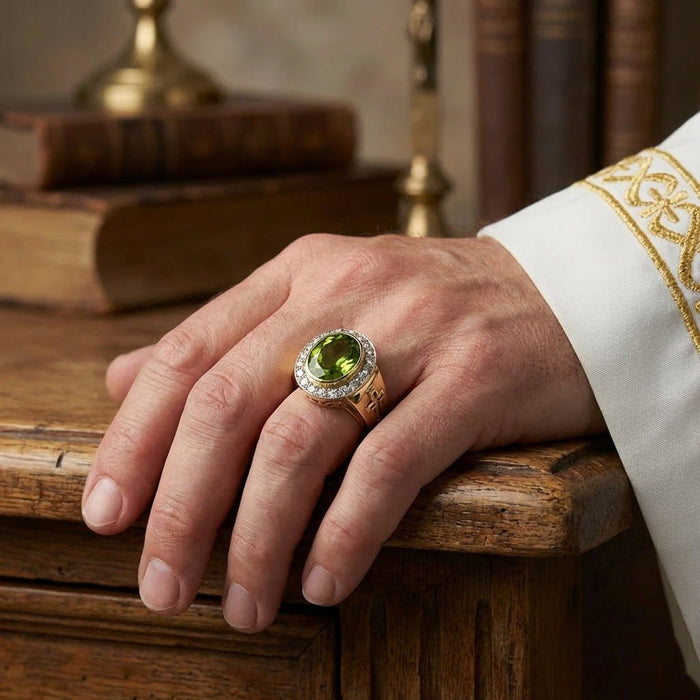 Hand resting on rustic wooden table wearing oval peridot bishop ring with CZ halo, gold embroidered white vestment cuff visible
