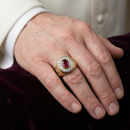 Yellow gold bishop ring with oval red ruby center stone surrounded by diamond halo worn on a man's ring finger resting on deep burgundy velvet fabric